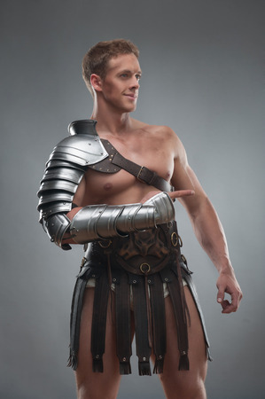 Half length portrait of young handsome muscular man gladiator in armour pointing aside isolated over grey backgroundの写真素材