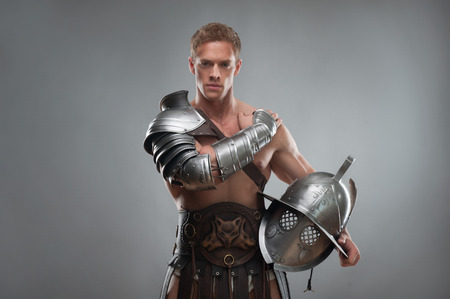 Half length portrait of young handsome muscular man gladiator in armour posing with helmet isolated over grey backgroundの写真素材