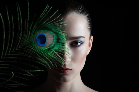 Beautiful brunette young woman with peacock style make-up and peacock feather isolated on black backgroundの写真素材