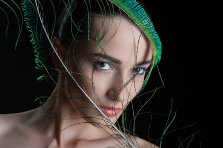 Beautiful brunette young woman with peacock style make-up and peacock feather, sideview  portrait isolated on black backgroundの写真素材