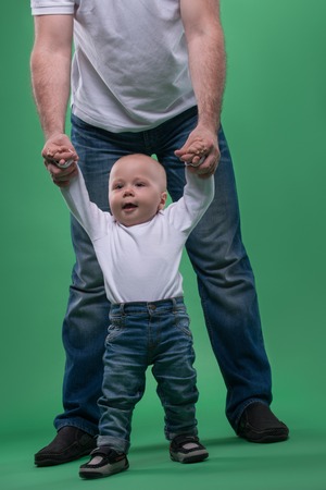 Portrait of little cute baby boy making first steps with his father, walking with support, isolated on greenの写真素材