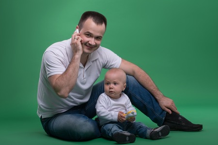 Happy handsome Caucasian father sitting on the floor with his son baby boy playing with toy cellphone, isolated on greenの写真素材
