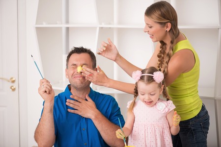 Family portrait, happy caucasian father, beautiful mother and daughter painting a picture with watercolours, handsome father having a yellow stain on his face on interior shotの写真素材