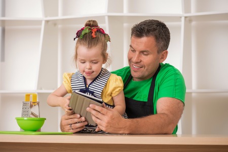 Half length portrait of handsome father and cute little daughter wearing aprons, cooking with tablet, grater and kitchen cutting board  Cooking or technology and food conceptの写真素材