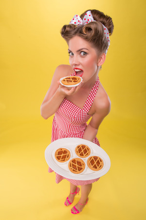 Top view portrait of beautiful coquette girl in dress with pretty smile in pinup style posing with tray of cookies and eating one cookie isolated on yellowの写真素材