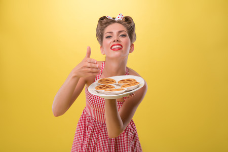 Half length portrait of beautiful coquette girl in dress with pretty smile in pinup style posing with tray of cookies isolated on yellowの写真素材