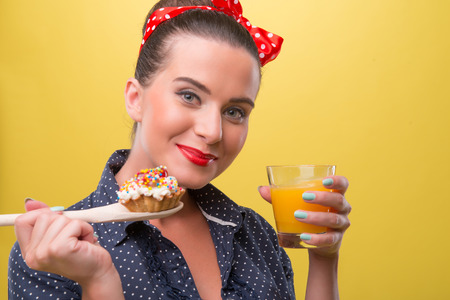 Half length clopseup portrait of beautiful sexy girl in polka dots dress with pretty smile in pinup style posing with cupcake and juice isolated on yellowの写真素材