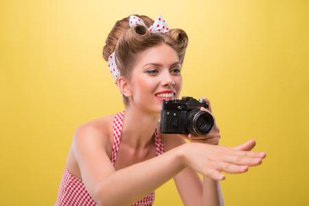 Half length portrait of beautiful sexy girl with pretty smile wearing dress in pin-up style posing with vintage photo camera isolated on yellowの写真素材