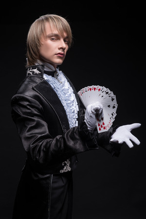 Half-length portrait of fair-haired serious juggler wearing interesting black costume and white shirt standing aside conjuring with the pack of cards in his hand  Isolated on black backgroundの写真素材