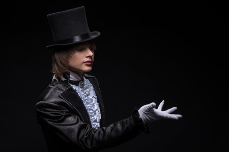 Half-length portrait of fair-haired matchless juggler wearing interesting black costume white shirt and topper standing aside showing us mastery of his hands  Isolated on black backgroundの写真素材