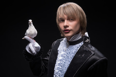 Half-length portrait of fair-haired matchless juggler wearing interesting black costume and white shirt holding pouter on his hand thinking about the next trick  Isolated on black backgroundの写真素材