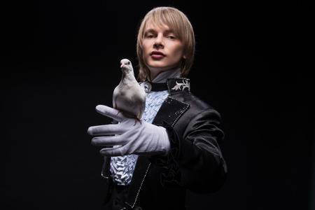 Half-length portrait of fair-haired matchless juggler wearing interesting black costume and white shirt showing us his good friend  Isolated on black backgroundの写真素材