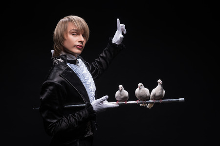 Half-length portrait of fair-haired matchless juggler wearing interesting black costume and white shirt standing aside holding three pouters on the silver stick  Isolated on black backgroundの写真素材