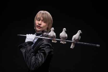 Half-length portrait of fair-haired satisfied juggler wearing interesting black costume and white shirt holding three pouters on the silver stick on his shoulder  Isolated on black backgroundの写真素材