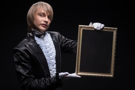 Half-length portrait of fair-haired matchless juggler wearing interesting black costume and white shirt showing us mysterious trick with the empty frame  Isolated on black backgroundの写真素材