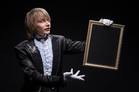 Half-length portrait of fair-haired matchless juggler wearing interesting black costume and white shirt showing us mysterious trick with the empty frame  Isolated on black backgroundの写真素材