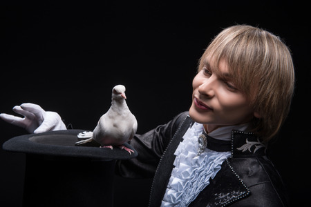 Half-length portrait of fair-haired smiling matchless juggler wearing interesting black costume and white shirt showing us the trick with the topper and pouter in it  Isolated on black backgroundの写真素材