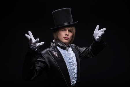 Half-length portrait of fair-haired matchless juggler wearing interesting black costume and white shirt standing with great black top on his head invited us to visit his performance  Isolated on black backgroundの写真素材