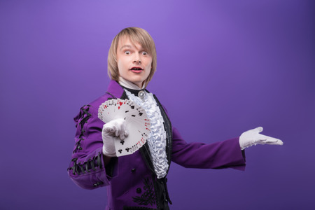 Half-length portrait of fair-haired excited juggler wearing splendid violet costume and white shirt showing us his great fen made of cards  Isolated on blue backgroundの写真素材