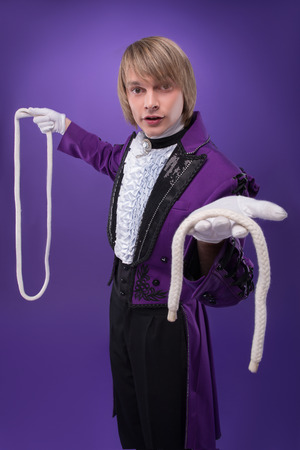 Half-length portrait of fair-haired concentrated juggler wearing splendid violet jacket and white shirt showing us two halves of the white rope  Isolated on black backgroundの写真素材