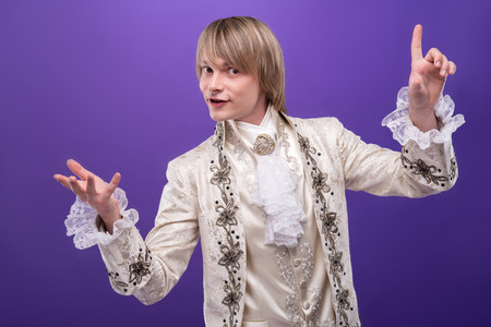 Half-length portrait of handsome fair-haired juggler wearing great white costume opening us the secret of his tricks  Isolated on violet backgroundの写真素材