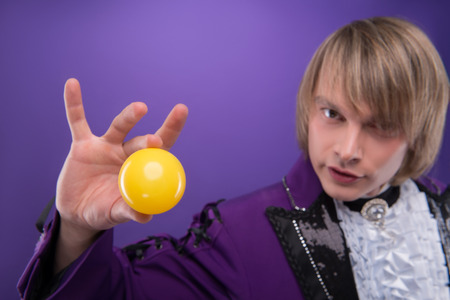 Selected focus on the small yellow ball in the hand of fair-haired enigmatic juggler wearing splendid violet jacket on backgroundの写真素材