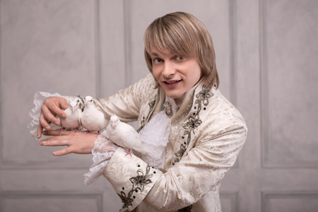 Half-length portrait of fair-haired smiling juggler wearing great white costume holding three white pouters on his hand satisfied with his performanceの写真素材