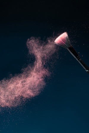 Vertical close-up photo of professional   black make-up brush with pink powder in motion isolated on blue background with copy placeの写真素材