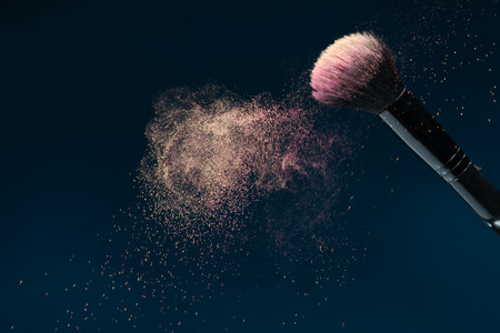 Horizontal close-up photo of professional   black make-up brush with pink powder in motion isolated on blue background with copy placeの写真素材