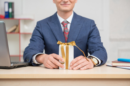 White present box with gold ribbons standing on the table in hands with a clock of young businessman in office surrounded by computer,notebook and mobilephoneの写真素材