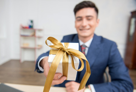 Close-up selective focus on a gift box in hands of young businessman in office working on his computer,showing a box to the cameraの写真素材