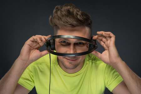Close-up portrait of smiling young DJ with stylish haircut holding looking through his headphone standing isolated over dark background の写真素材