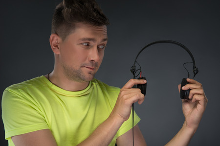 Close-up portrait of smiling young DJ with stylish haircut holding and looking his headphone standing isolated over dark background の写真素材