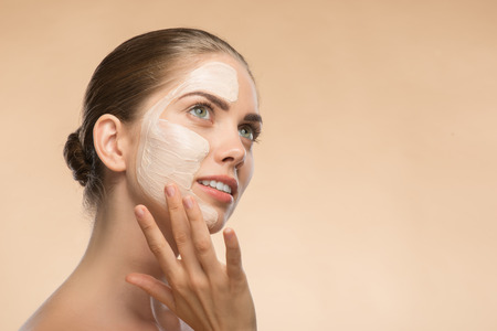 Horizontal Portrait of beautiful dreamy girl in spa with cosmetic cream on her face with clean and  fresh skin looking aside and touching her face isolated on beige background with copy placeの写真素材