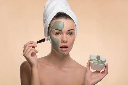 Portrait of surprised looking at the camera girl in spa with a  towel on her head applying facial clay mask with brush on her face and holding a plate isolated on beige background with copy placeの写真素材