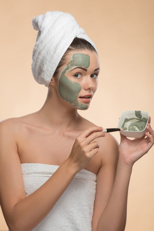 Waist-up portrait of beautiful looking at the camera girl in spa with a  towel on her head applying facial clay mask with brush on her face and holding a plate isolated on beige backgroundの写真素材