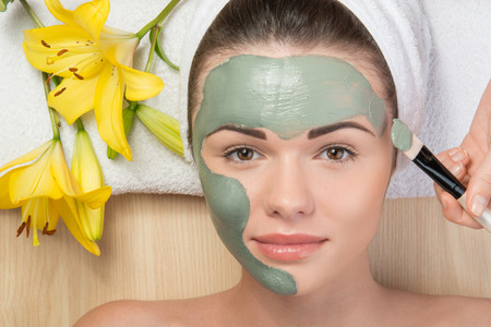 Close-up portrait of beautiful girl looking at the camera with a towel on her head applying facial clay mask and beauty treatments lying on a table in spa near yellow flowerの写真素材