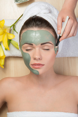 Close-up portrait of beautiful girl with closed eyes  with a towel on her head applying facial clay mask and beauty treatments lying on a table in spa near yellow flowerの写真素材