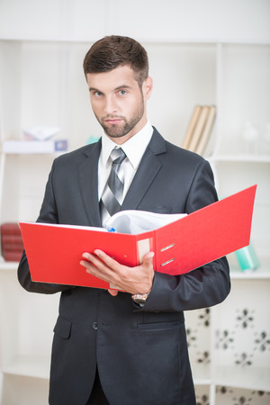 Waist-up portrait of handsome confident businessman standing in office with a red folder in his hands and looking at the camera with serious faceの写真素材