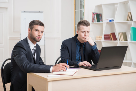Portrait of Two handsome businessmen in office working on some project, one stressed looking at laptop and another writing in his notebook and looking at the cameraの写真素材
