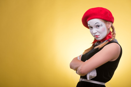 Close-up portrait of indignant female mime with white funny face, red hat and red scarf looking at the camera isolated on yellow background with copy placeの写真素材