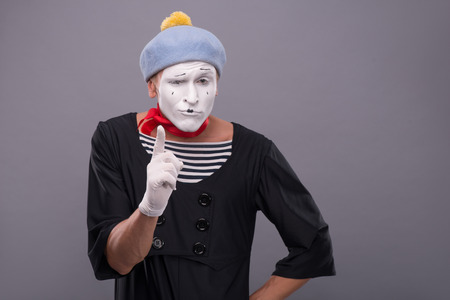 Waist-up portrait of funny male mime with grey hat and white face showing sign Attention and looking at the camera isolated on grey backgroundの写真素材