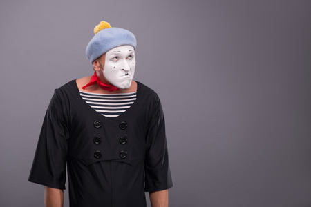 Waist-up portrait of funny male mime with grey hat and white face disappointed looking up and holding his hands omitted down isolated on grey background with copy placeの写真素材