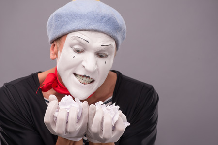 Close-up portrait of crazy male mime with grey hat and white face holding crumped paper and looking at this paper with interest isolated on grey backgroundの写真素材
