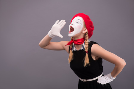 Waist-up portrait of funny female mime in red head and with white face very loudly shouting and looking aside isolated on grey background with copy placeの写真素材
