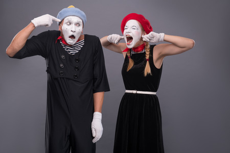 Waist-up portrait of mime couple with white faces, female mime closing her ears with her fingers and shouting on male mime showing that she is crazy isolated on grey backgroundの写真素材
