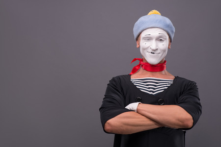 Waist-up portrait of male mime with grey hat and white face funny smiling to the camera and  holding hands on his chest isolated on grey background with copy placeの写真素材
