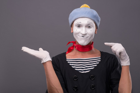Close-up portrait of male mime with grey hat and white face funny smiling to the camera and showing with finger on his left hand isolated on grey backgroundの写真素材