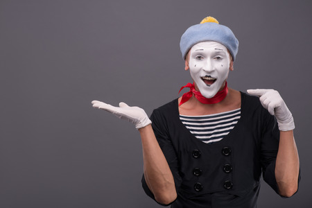 Close-up portrait of male mime with grey hat and white face funny smiling to the camera and showing with finger on his left hand isolated on grey backgroundの写真素材
