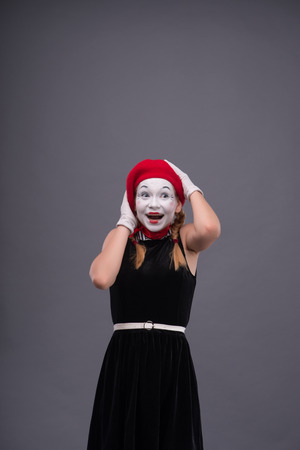 portrait of female mime with surprised face, red hat and red scarf looking aside smiling and touching her head isolated on grey backgroundの写真素材